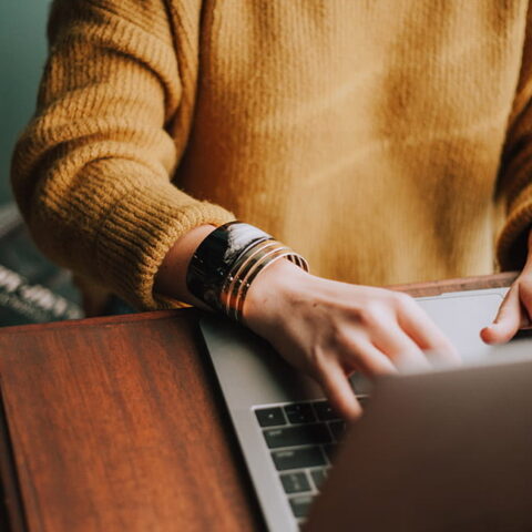 A person in a yellow sweater types on a laptop. Their face is not shown, just their torso and hands.
